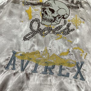 May include: A satin bomber jacket with embroidered details. The design features the word "BAHAMA" above a skull, the name "Joe's", a compass, and the word "AVIREX". The embroidery is in shades of gray, yellow, and brown.