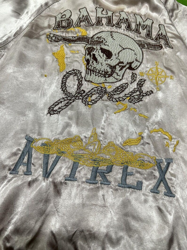 May include: A satin bomber jacket with embroidered details. The design features the word "BAHAMA" above a skull, the name "Joe's", a compass, and the word "AVIREX". The embroidery is in shades of gray, yellow, and brown.