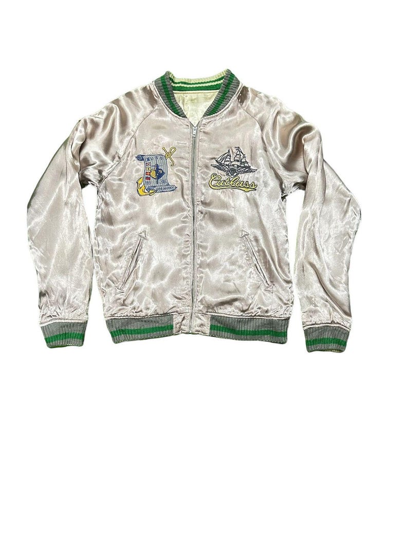 May include: A light silver satin bomber jacket with green and gray striped trim. The jacket features embroidered patches including a ship and the word "Cutlass". The jacket has a zipper closure and two front pockets.