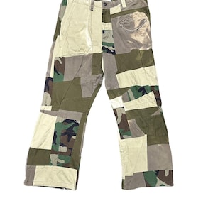 Op de afbeelding: Patchworkbroek in diverse kleuren, waaronder olijfgroen, beige en camouflagepatronen. De broek heeft een knoop- en ritssluiting, riemlussen en meerdere zakken. Het ontwerp bevat een mix van stoffen en texturen.