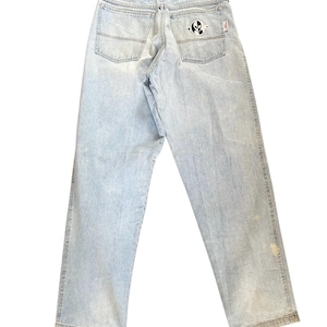 May include: Light blue denim jeans with a straight leg cut. The back pockets feature a black and white logo. The jeans have a classic five-pocket design and a button and zipper closure. The denim has a faded wash and some light discoloration.