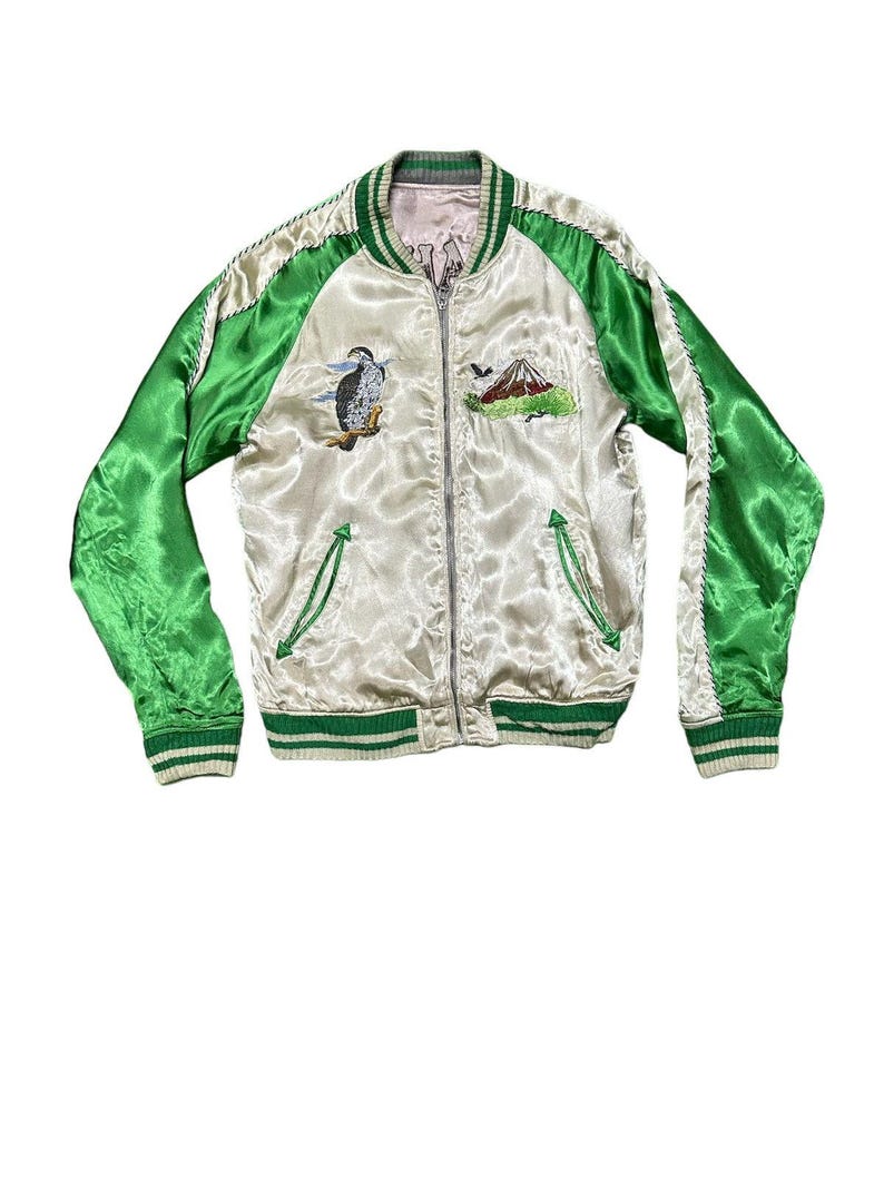 May include: Cream-colored satin bomber jacket with green sleeves and trim. Features embroidered images of a bird and a mountain. The jacket has a zipper closure and two front pockets.