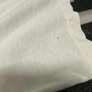 May include: Close-up of a white t-shirt, highlighting the fabric's texture. The shirt is slightly creased, with a small flaw visible. The image focuses on the material and construction of the garment.
