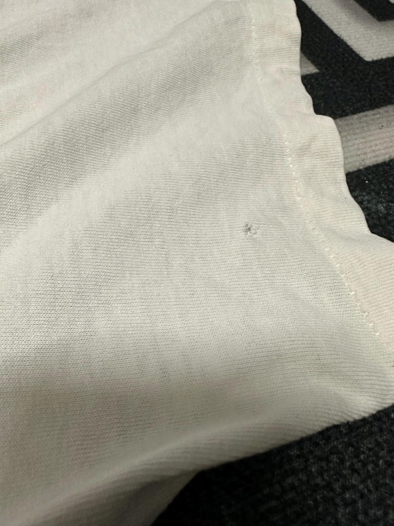 May include: Close-up of a white t-shirt, highlighting the fabric's texture. The shirt is slightly creased, with a small flaw visible. The image focuses on the material and construction of the garment.
