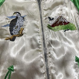 May include: Cream-colored satin bomber jacket with green trim and a silver zipper. The jacket features embroidered designs of a hawk perched on a branch, a mountain, and a flying bird. The jacket has a ribbed collar and cuffs.