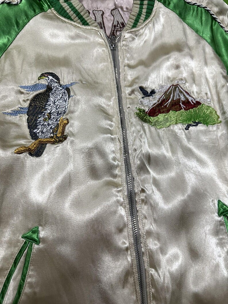 May include: Cream-colored satin bomber jacket with green trim and a silver zipper. The jacket features embroidered designs of a hawk perched on a branch, a mountain, and a flying bird. The jacket has a ribbed collar and cuffs.