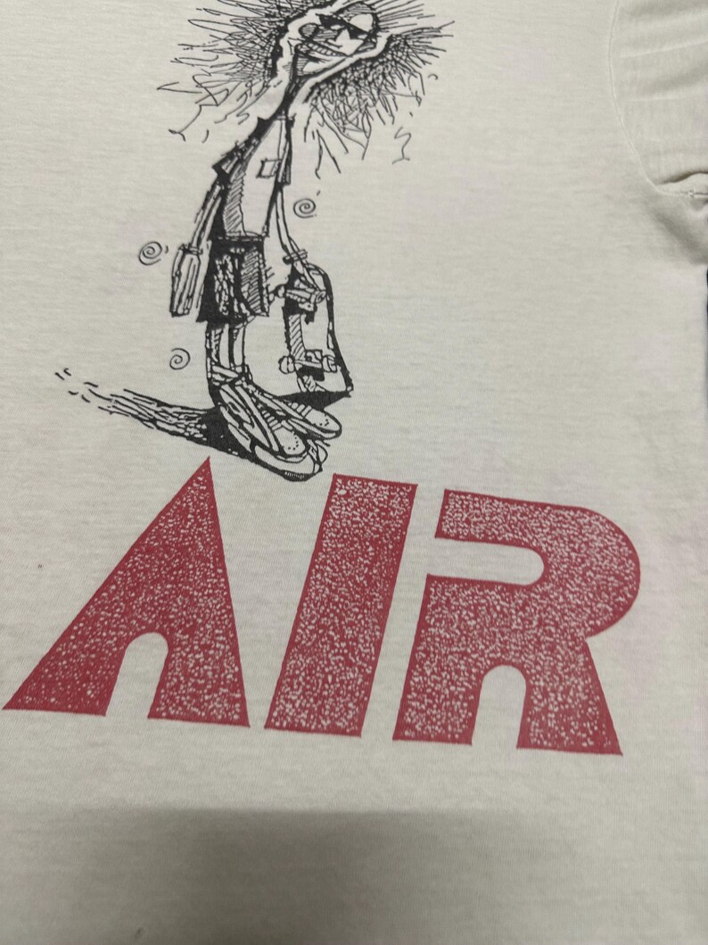 May include: White t-shirt with a cartoon skater graphic in black and white, and the word "AIR" in large red letters. The skater is drawn in a whimsical style, holding a skateboard.