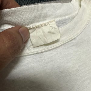 May include: Close-up of a white t-shirt collar and label. The label is a light beige colour and appears to be made of fabric. The t-shirt is made of a soft, textured material.