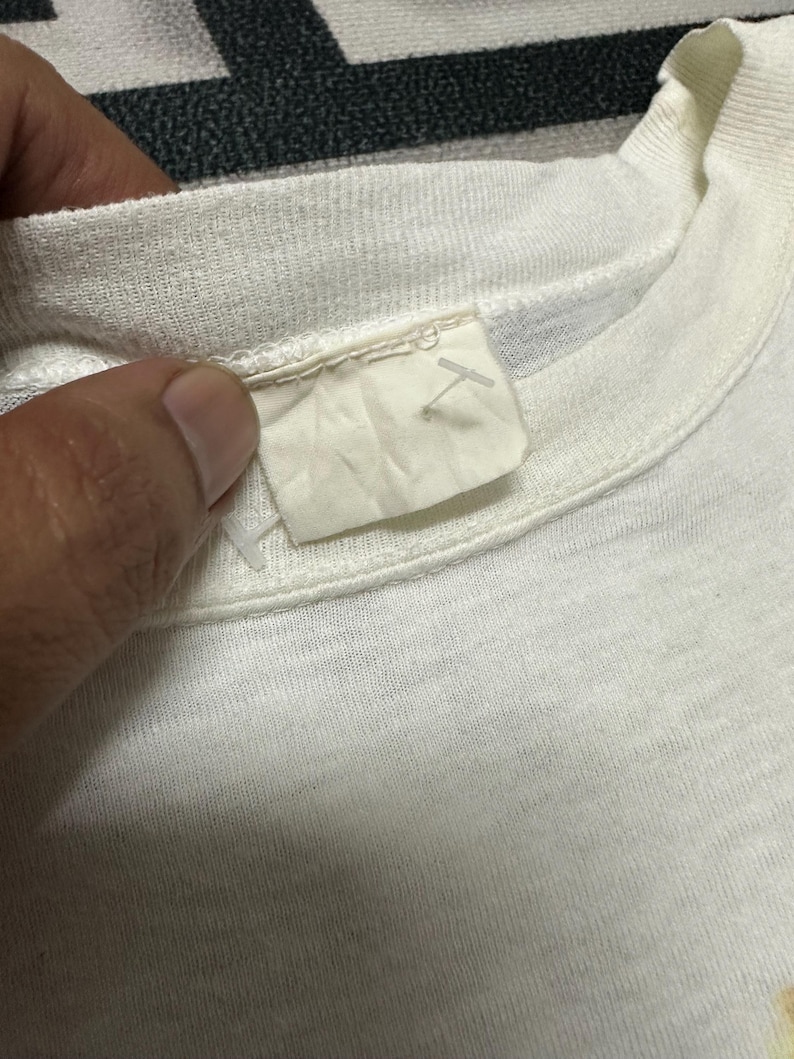 May include: Close-up of a white t-shirt collar and label. The label is a light beige colour and appears to be made of fabric. The t-shirt is made of a soft, textured material.