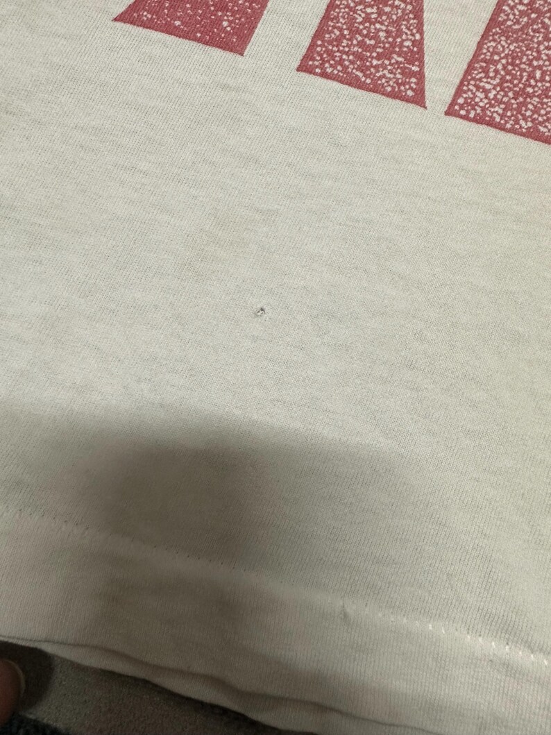 May include: Close-up of a light-coloured t-shirt with a red, distressed text design at the top. The fabric appears to be a soft cotton blend. The bottom hem is visible.