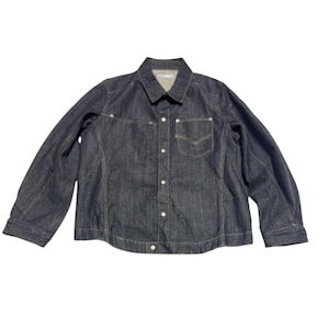 Puede incluir: Chaqueta vaquera azul oscuro con cierre de botones, bolsillo en el pecho y cuello. La chaqueta tiene mangas largas y un diseño clásico. Una pequeña etiqueta roja es visible en el bolsillo.