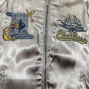 May include: A light gray satin bomber jacket with a full zipper closure. The jacket features embroidered patches including a nautical design with the text "USN" and "USS AVIREX", and a ship with the word "Cutlass". The collar has green and gray stripes.