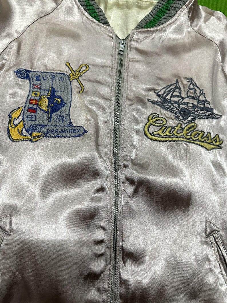 May include: A light gray satin bomber jacket with a full zipper closure. The jacket features embroidered patches including a nautical design with the text "USN" and "USS AVIREX", and a ship with the word "Cutlass". The collar has green and gray stripes.