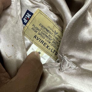 May include: Close-up of a tan satin jacket lining with a label that reads "EQUIPMENT-TYPE-A" and "AVIREX LTD". The label also includes text about flight garment specifications and a stock number. The jacket is a size M.