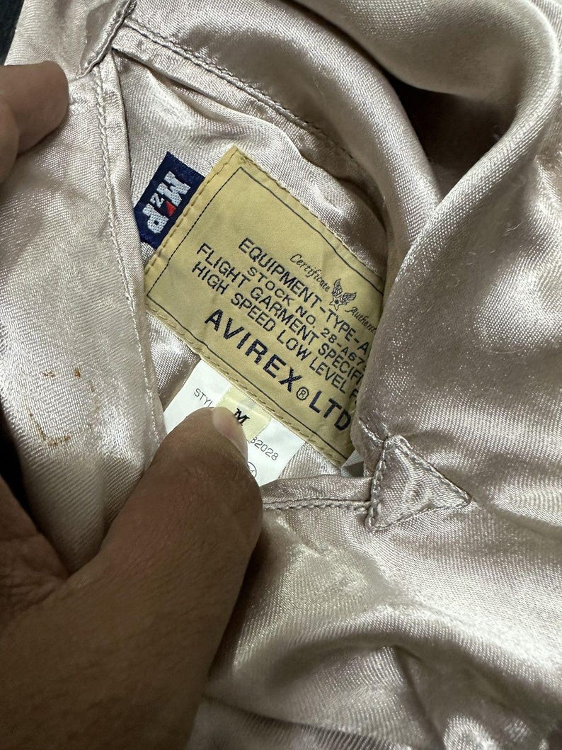 May include: Close-up of a tan satin jacket lining with a label that reads "EQUIPMENT-TYPE-A" and "AVIREX LTD". The label also includes text about flight garment specifications and a stock number. The jacket is a size M.