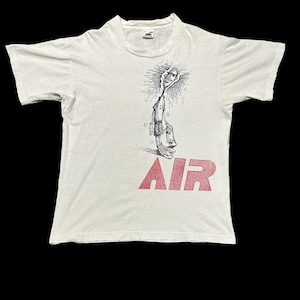 May include: Cream-coloured t-shirt with a black and white illustration of a figure and the word "AIR" in red block letters. The tee has short sleeves and a crew neck.