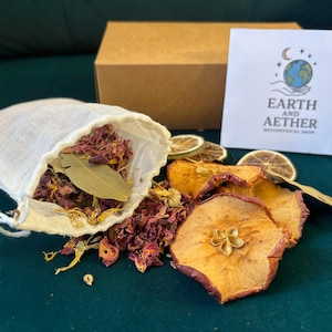 Può includere: Un sacchetto di mussola bianca pieno di petali di rosa secchi, foglie di alloro e altri prodotti botanici, insieme a fette di mela essiccate e una carta con il testo "Earth and Aether Metaphysical Shop". Una scatola marrone è sullo sfondo.