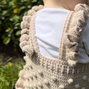 Puede incluir: Un pelele de ganchillo beige con bolitas texturizadas y bordes con volantes. Las correas y el escote presentan un patr&oacute;n de ganchillo detallado. La prenda se lleva sobre una camiseta blanca, lo que sugiere un atuendo para beb&eacute; o ni&ntilde;o peque&ntilde;o.