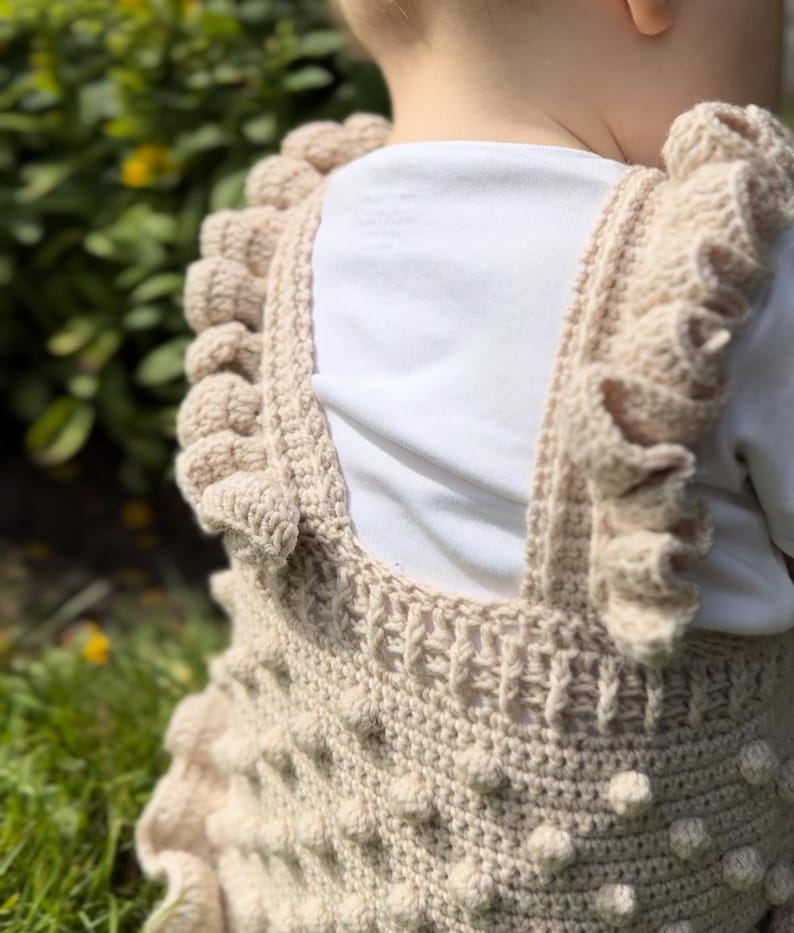 Puede incluir: Un pelele de ganchillo beige con bolitas texturizadas y bordes con volantes. Las correas y el escote presentan un patr&oacute;n de ganchillo detallado. La prenda se lleva sobre una camiseta blanca, lo que sugiere un atuendo para beb&eacute; o ni&ntilde;o peque&ntilde;o.