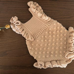 Puede incluir: Un pelele de ganchillo hecho a mano de color beige claro, con bordes y tirantes con volantes. El pelele presenta un patr&oacute;n de puntadas texturizadas y est&aacute; dise&ntilde;ado para un beb&eacute; o un ni&ntilde;o peque&ntilde;o. La prenda se exhibe sobre una superficie de madera marr&oacute;n oscuro.
