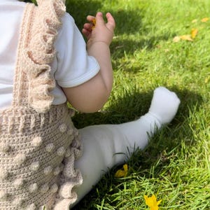 Puede incluir: Un beb&eacute; con un pelele de ganchillo beige, una camiseta blanca y medias blancas, sentado en hierba verde. El pelele tiene un dise&ntilde;o texturizado con bolitas y bordes con volantes. El beb&eacute; sostiene una flor amarilla.