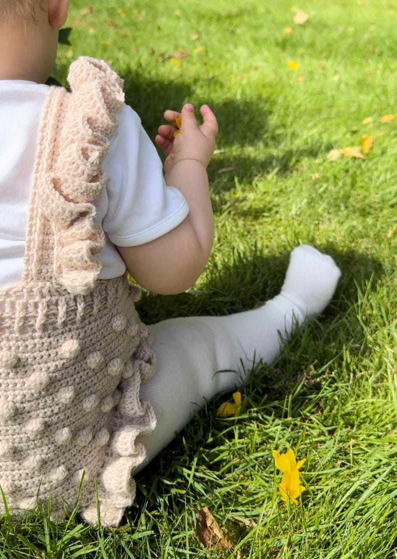 Puede incluir: Un beb&eacute; con un pelele de ganchillo beige, una camiseta blanca y medias blancas, sentado en hierba verde. El pelele tiene un dise&ntilde;o texturizado con bolitas y bordes con volantes. El beb&eacute; sostiene una flor amarilla.