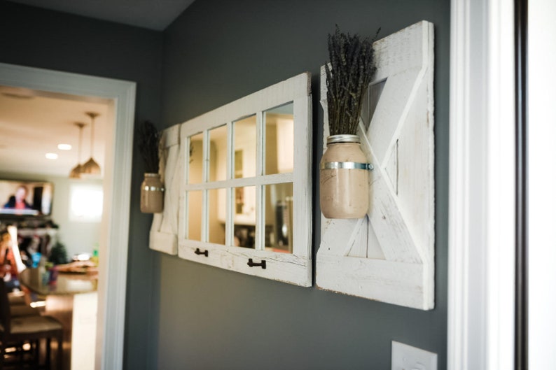 May include: Three white painted wooden wall decor pieces with a farmhouse style. The center piece is a mirror with a windowpane design. The pieces on either side are a rustic barn door design with a mason jar holding dried lavender flowers.