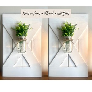 May include: Two white wooden shutters with a mason jar and greenery in each. The mason jars are clear glass with a white lid and a twine bow. The greenery is a small bush of green leaves. The text "Mason Jars + Floral + Shutters" is written in black on a tan background at the top of the image.