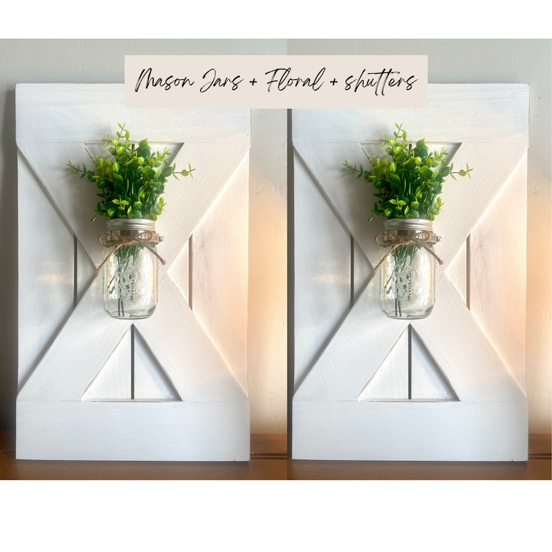 May include: Two white wooden shutters with a mason jar and greenery in each. The mason jars are clear glass with a white lid and a twine bow. The greenery is a small bush of green leaves. The text "Mason Jars + Floral + Shutters" is written in black on a tan background at the top of the image.