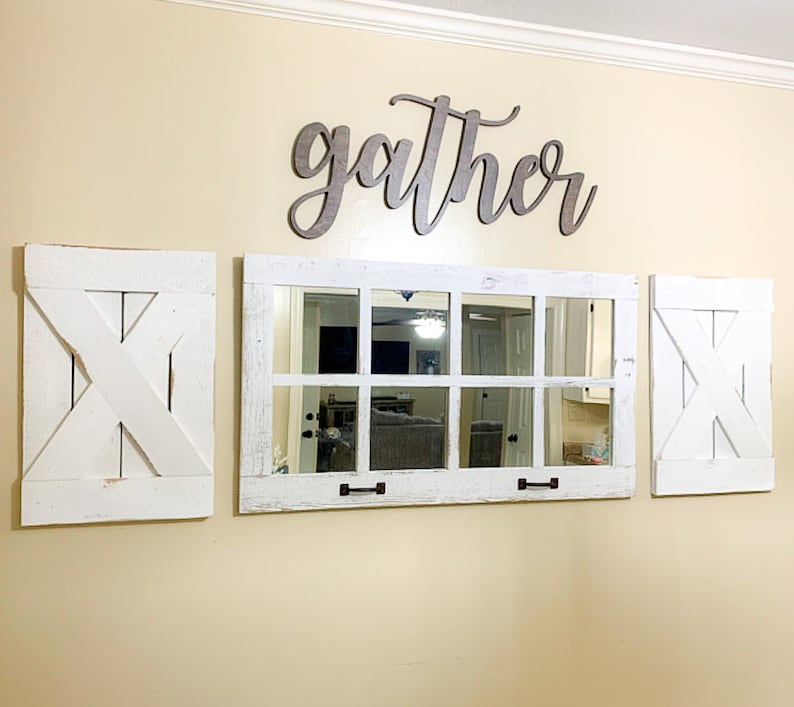 May include: A white farmhouse style mirror with a wood frame and a distressed finish. The mirror is divided into nine squares and has two black metal handles. The mirror is flanked by two white shutters with a criss-cross design. The word "gather" is written in cursive above the mirror.