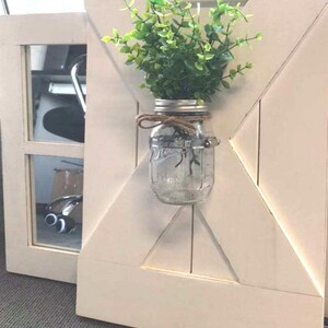 May include: A white wooden door with a decorative X-shaped design. A mason jar with a green plant is attached to the door.