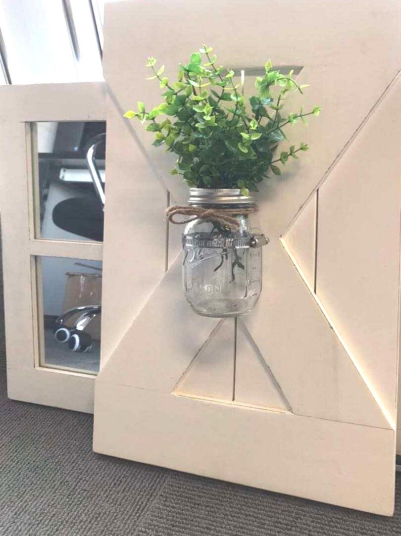 May include: A white wooden door with a decorative X-shaped design. A mason jar with a green plant is attached to the door.