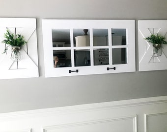 May include: Three white framed mirrors with a farmhouse style design. The center mirror is larger and has nine panes of glass. The two smaller mirrors are on either side of the center mirror and have a single pane of glass. Each mirror has a small potted plant in a glass jar.