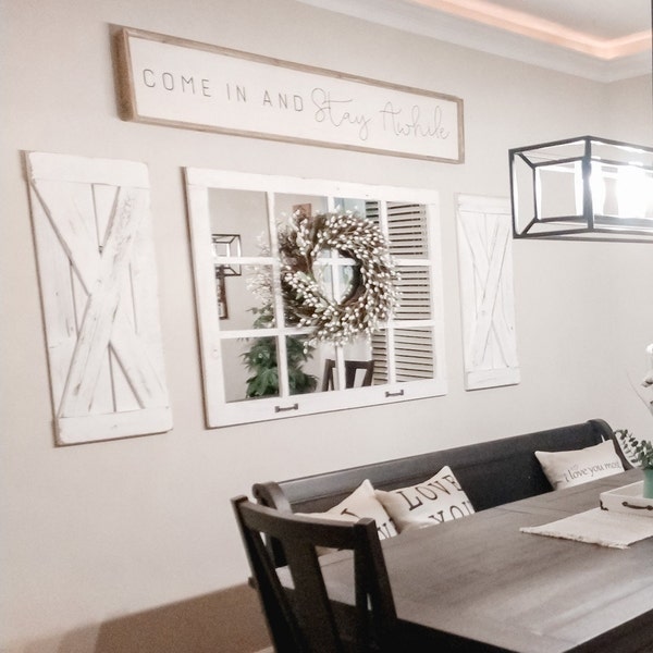 May include: A dining room with a farmhouse style. The walls are painted a light beige color. There is a wooden sign that reads "Come in and Stay Awhile". There are two white shutters on the wall, a mirror with a wreath, and a black bench with pillows that read "Love You".