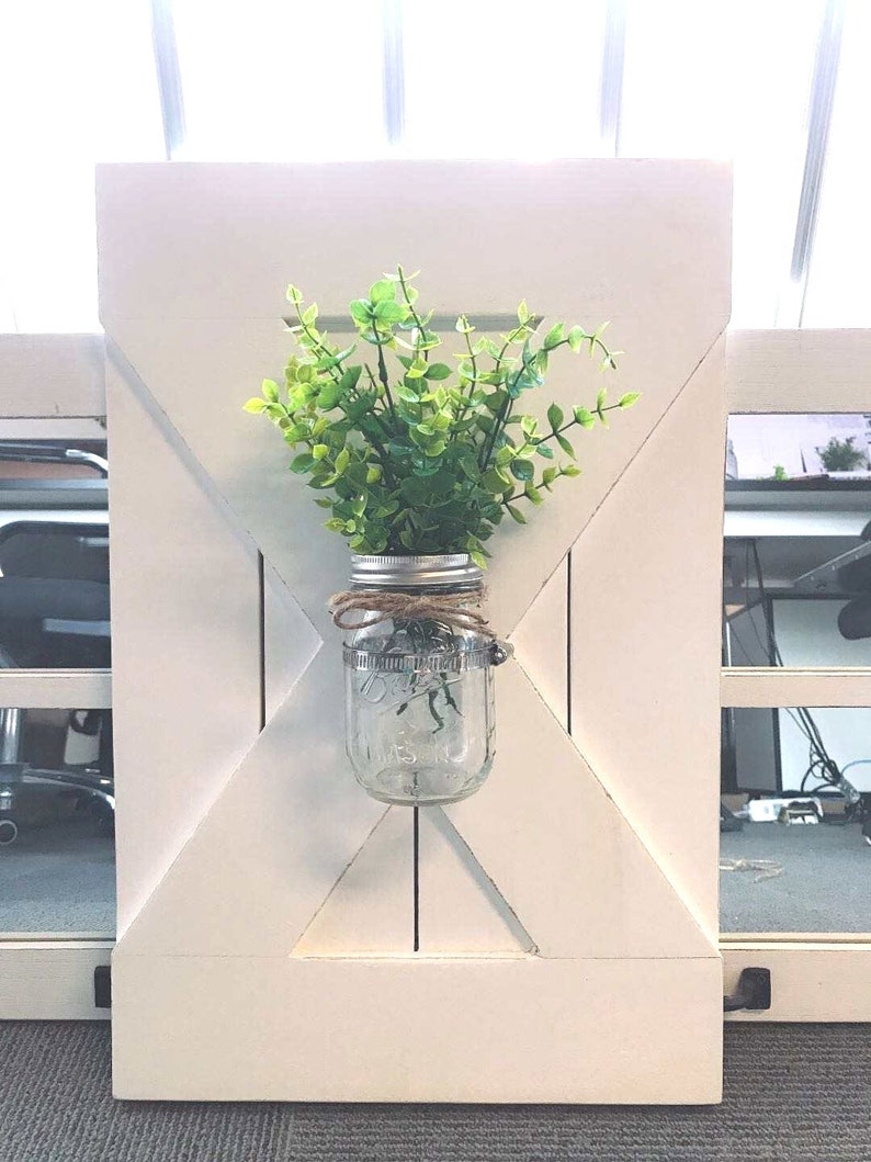 May include: White wooden farmhouse style wall decor with a mason jar filled with green faux greenery hanging in the centre.