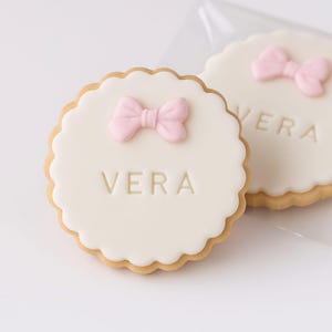 Op de afbeelding: Twee ronde koekjes met geschulpte randen. Elk koekje heeft een witte glazuurbasis met de naam "VERA" in reliëf in het midden. Op elk koekje is een roze strik geplaatst. De koekjes staan op een witte achtergrond.
