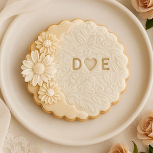 Peut inclure: Un biscuit au sucre décoré avec un motif floral et les lettres "D", un cœur et "E". Le biscuit est sur une assiette blanche, avec trois accents de fleurs et un bord festonné. Des roses roses et du tissu sont en arrière-plan.