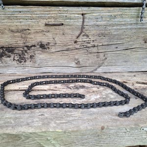 May include: A dark gray bicycle chain is arranged in a curved pattern on a weathered wooden surface. The chain's links are visible, and the background features aged wooden planks with visible grain and knots.