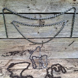 May include: Several metal chains of varying lengths and designs are displayed against a weathered wooden background. The chains are primarily silver and dark gray, with some appearing to be rusted. The arrangement includes straight and curved chain segments.