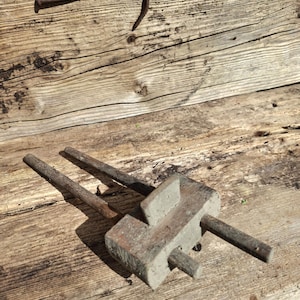 May include: An antique wooden marking gauge with two metal rods and a rectangular block. The tool rests on a weathered wooden surface, with two rusty nails in the background. The wood has visible grain and knots.