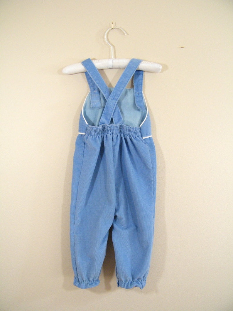 Op de afbeelding: Blauwe corduroy broek met witte bies en verstelbare schouderbanden. De broek hangt aan een witte hanger.