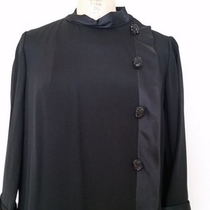 K&ouml;nnte beinhalten: Ein schwarzes, lang&auml;rmeliges Kleid mit einem Satin-Panel an der Seite und schwarzen Strass-Kn&ouml;pfen. Das Kleid befindet sich auf einer Kleiderpuppe.