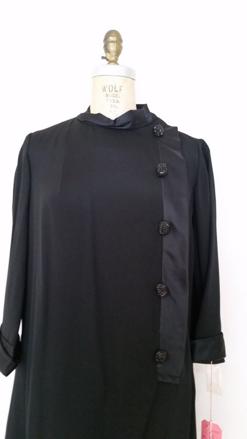 K&ouml;nnte beinhalten: Ein schwarzes, lang&auml;rmeliges Kleid mit einem Satin-Panel an der Seite und schwarzen Strass-Kn&ouml;pfen. Das Kleid befindet sich auf einer Kleiderpuppe.