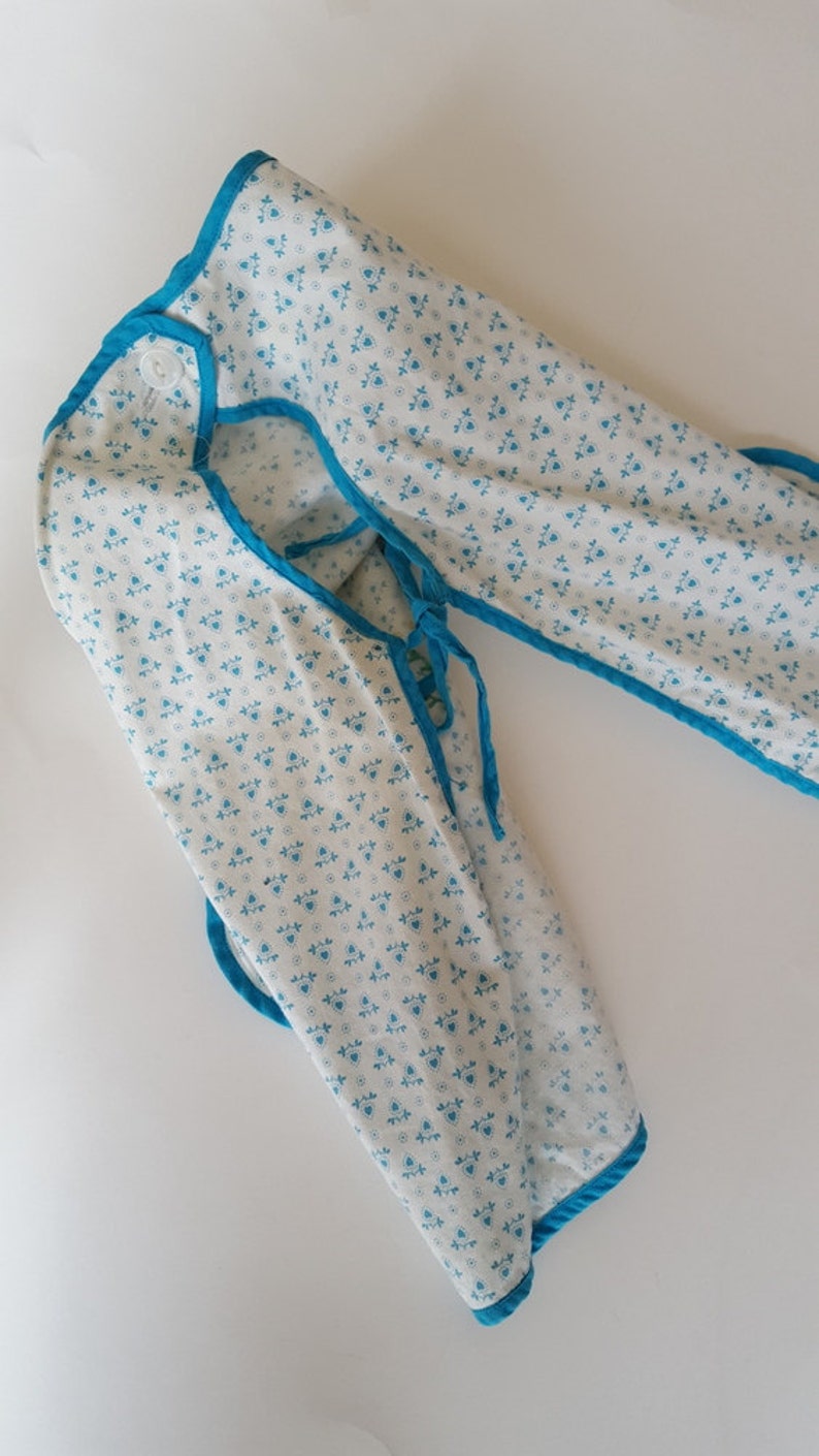 May include: A white fabric baby bib with a blue trim and a small white button. The bib has a blue and white floral pattern.