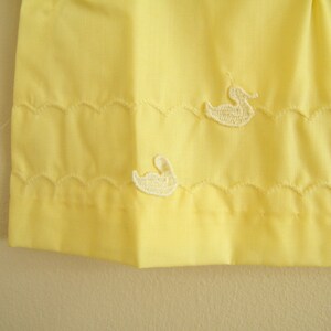 May include: A yellow fabric skirt with white embroidered ducks. The skirt has a scalloped hem.