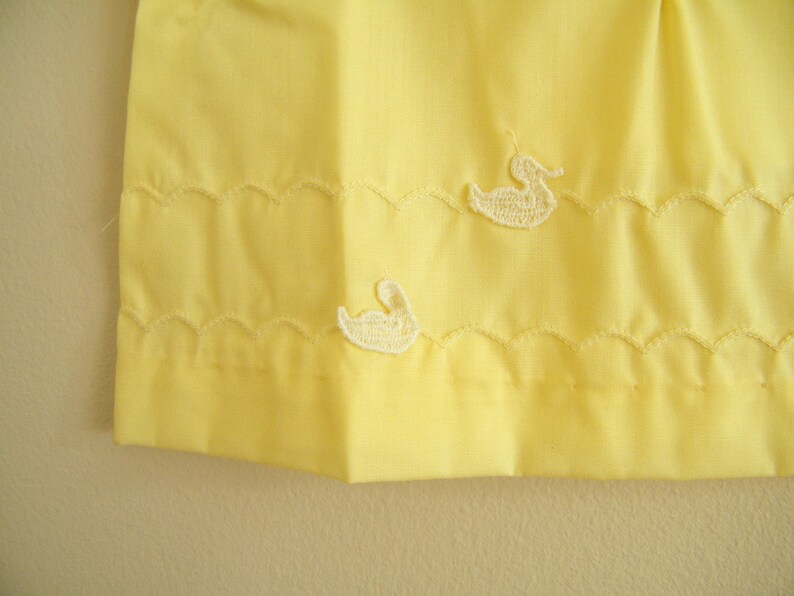 May include: A yellow fabric skirt with white embroidered ducks. The skirt has a scalloped hem.