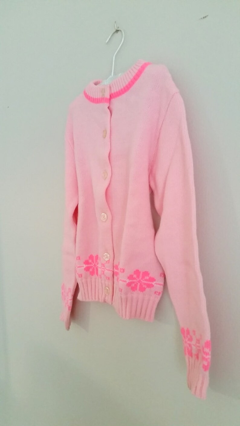 Puede incluir: Un c&aacute;rdigan de punto rosa con cierre de botones y un dise&ntilde;o floral rosa en el dobladillo inferior y los pu&ntilde;os.
