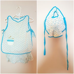 May include: A white and blue baby bib and matching romper with a floral pattern. The bib has a blue trim and ties. The romper has a blue trim and a pocket with a flower design.