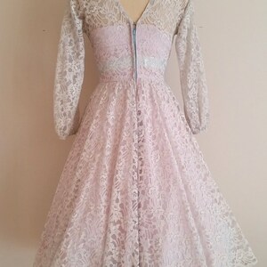 May include: A vintage, light pink lace dress with a V-neckline and long sleeves. The dress has a fitted bodice and a full skirt.