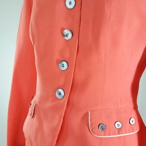 Puede incluir: Un traje vintage de color rosa coral con una chaqueta entallada y una falda. La chaqueta tiene un ribete blanco alrededor del cuello y los bolsillos. La chaqueta tiene cinco botones en la parte delantera, todos con un centro blanco y un anillo exterior gris oscuro.